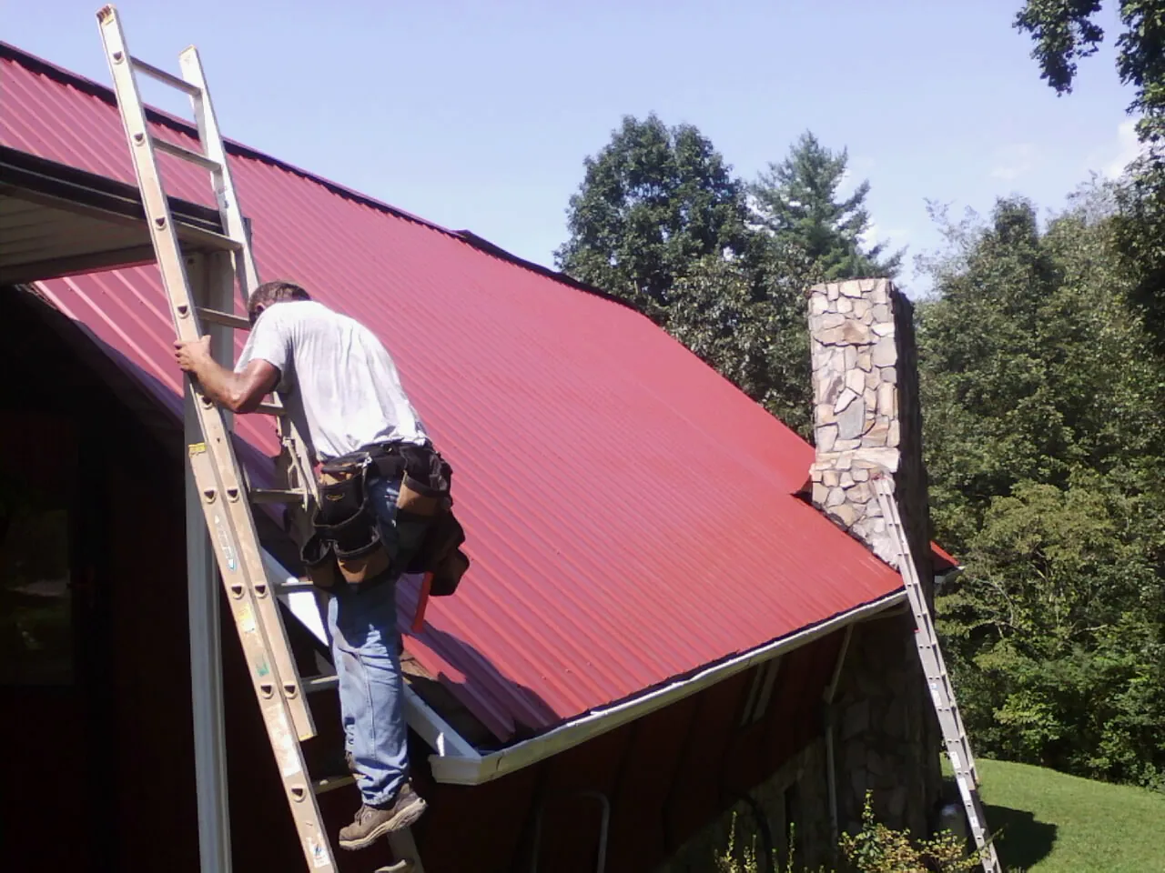 Roofer on a ladder beside a red metal roof and stone chimney — Soffit & Fascia Repair by Roofing Brothers in Montgomery